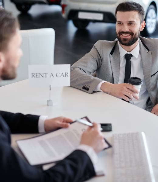 Smiling bearded businessman with coffee sitting at auto salon and communicating with salesman. He wants to rent a car