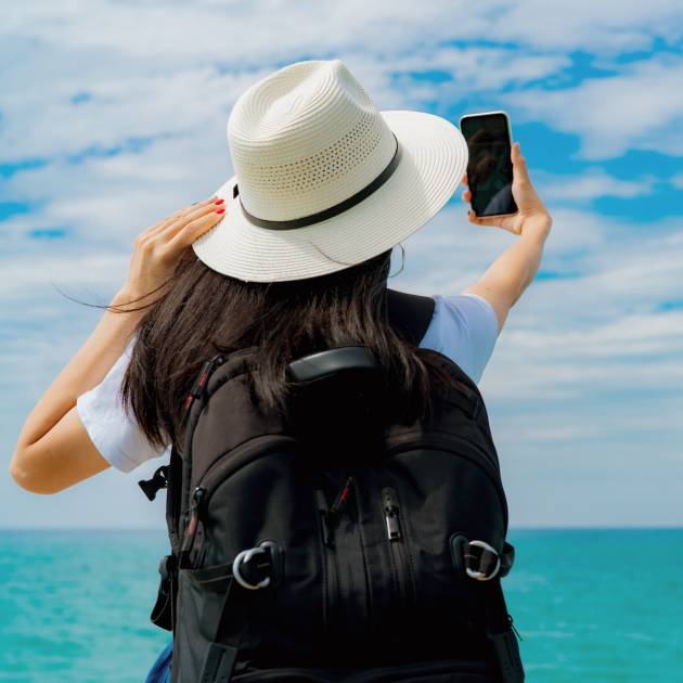Young Asian backpacker woman wear hat use smartphone taking selfie at pier. Summer vacation at tropical paradise beach. Happy hipster girl travel on holiday. Woman enjoy and relax life. Summer vibes.