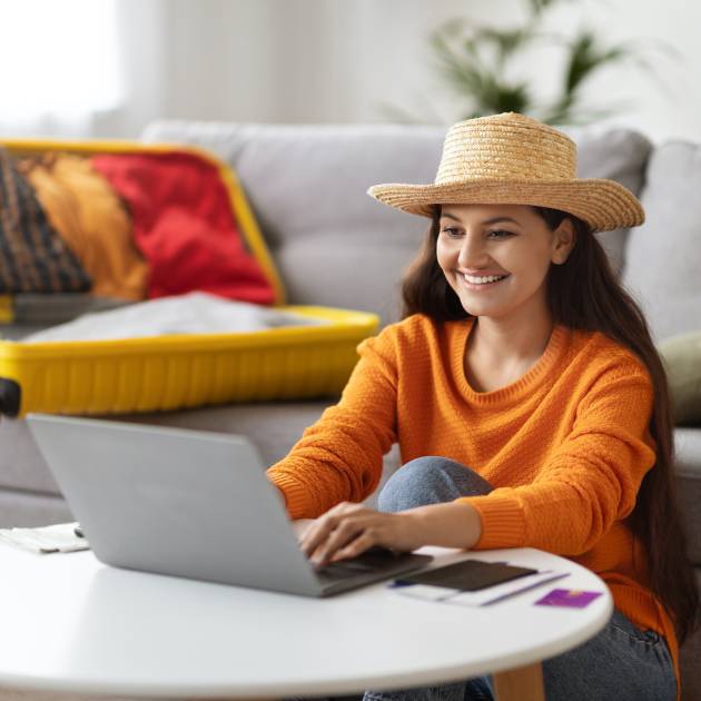 Excited happy young pretty indian woman wearing straw hat using laptop at home, packing suitcase, booking hotel or flight tickets online, planning trip, going vacation, copy space